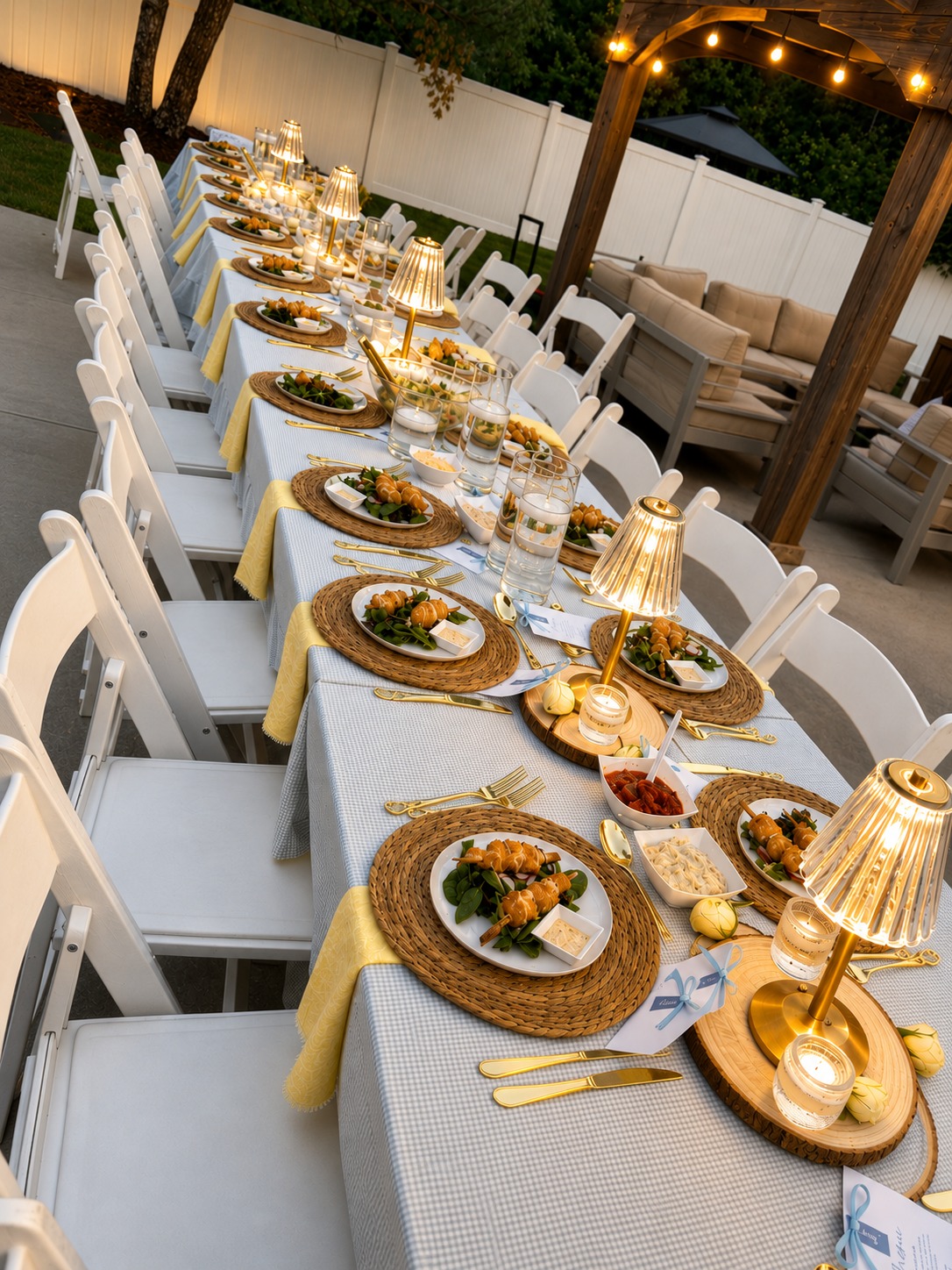 Outdoor summer dinner party fully set at dusk — long linen table with woven placemats, glowing lamps, and yellow accents